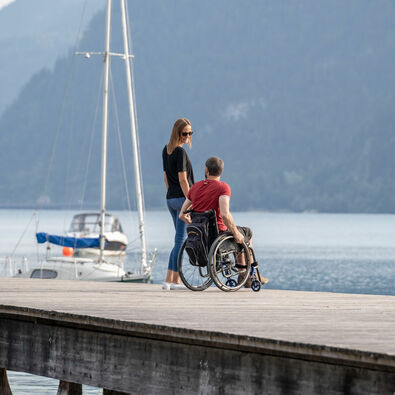 Ein Mann im Rollstuhl und eine Frau befinden sich auf dem Steg bei der Seeuferpromenade in Pertisau im Sommer. 