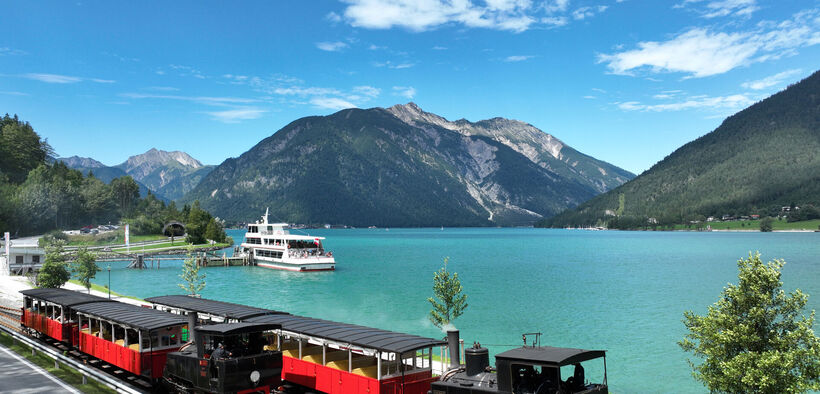 Die Luftaufnahme zeigt ein Schiff der Achenseeschifffahrt sowie die Achenseebahn an der Anlegestelle Seespitz im Sommer.