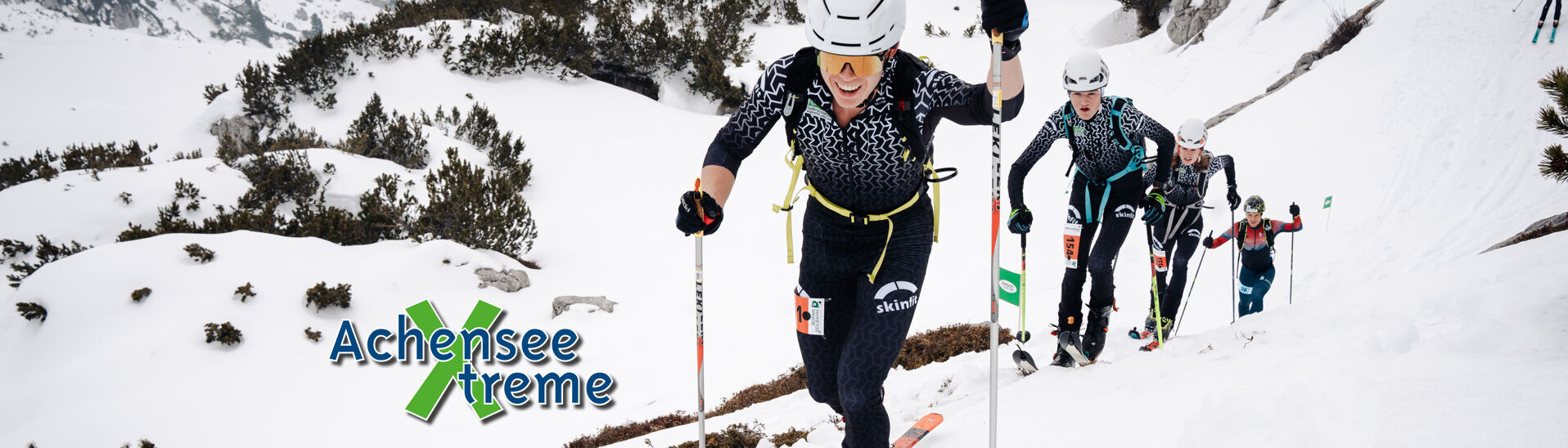 Achensee Xtreme Eine Gruppe von Skitourengehern erklimmt einen schneebedeckten Hang. Die Athleten tragen spezielle Skiausrüstung und konzentrieren sich auf den Aufstieg. Im Vordergrund ist ein Teilnehmer sichtbar, der lächelt und Stöcke zum Vorankommen nutzt. Im Hintergrund sind weitere Skifahrer zu sehen.}
