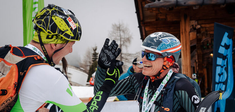 Two ski mountaineers celebrate their participation in the Achensee Xtreme Individual. One skier wears a black and gray outfit with a helmet, while the other wears a white and green outfit. Snowy surroundings and banners hint at a skiing event.