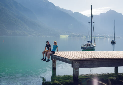 A couple exploring the lakeside promenade and the wooden pier in Pertisau am Achensee, backdropped by the Ebner Joch and several sailboats.