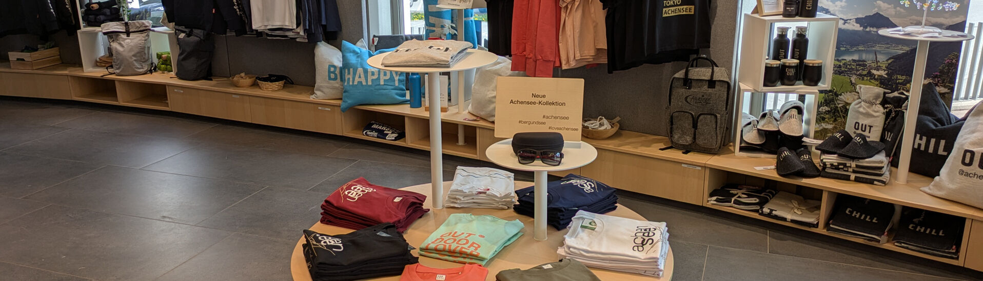 Ein Schild mit der Aufschrift „Neue Achensee-Kollektion“ steht neben mehreren T-Shirts in verschiedenen Farben. Auf einem weißen Shirt ist der Schriftzug „achensee“ zu sehen. Die Umgebung wirkt einladend und geschäftig, mit weiteren Kleidungsstücken im Hintergrund.}