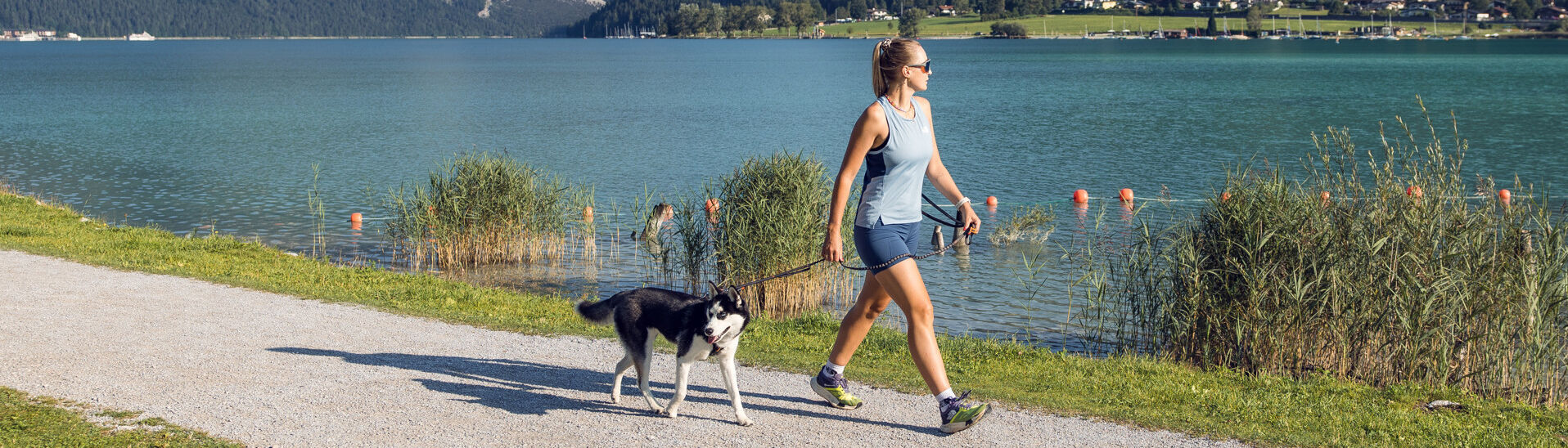 Hunde am Achensee Eine junge Frau geht mit ihrem Hund an der Leine am Ufer des Achensees spazieren. Im Hintergrund sind Berge und eine grüne Landschaft zu sehen. Es ist ein schöner, klarer Tag.}