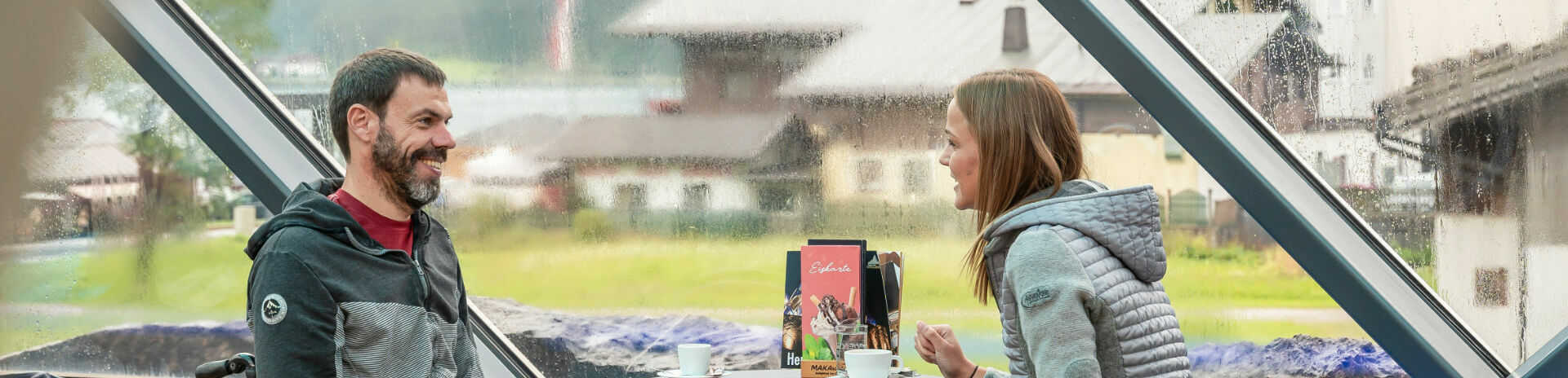 Zwei Freunde bei denen einer im Rollstuhl sitzt unterhalten sich im Cafe Vitalberg mit einem heißen Getränk bei Regenwetter.}