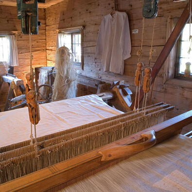 Ein traditioneller Webstuhl steht in einem Holzraum. Der Tisch des Webstuhls ist mit einem weißen Tuch bedeckt. Im Hintergrund sind mehrere Spinnräder und Arbeitswerkzeuge zu sehen. Natürliches Licht fällt durch die Fenster und beleuchtet den Raum sanft.
