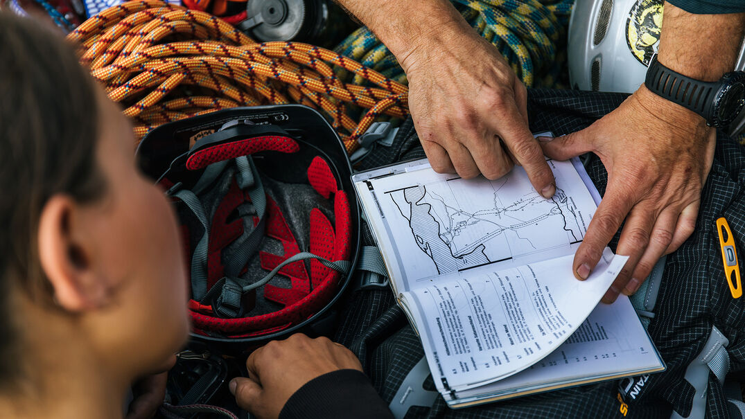 Ein Mann der einer Frau gerade eine Karte erklärt, welche neben einem Kletterhelm und einem Seil liegt.