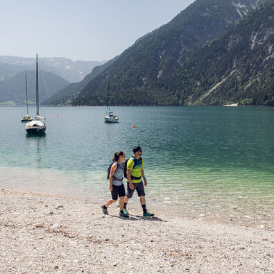 Ein Paar genießt einen kleinen Spaziergang am Ufer des Achensees in Achenkirch. Im Hintergrund sieht man Segelboote.