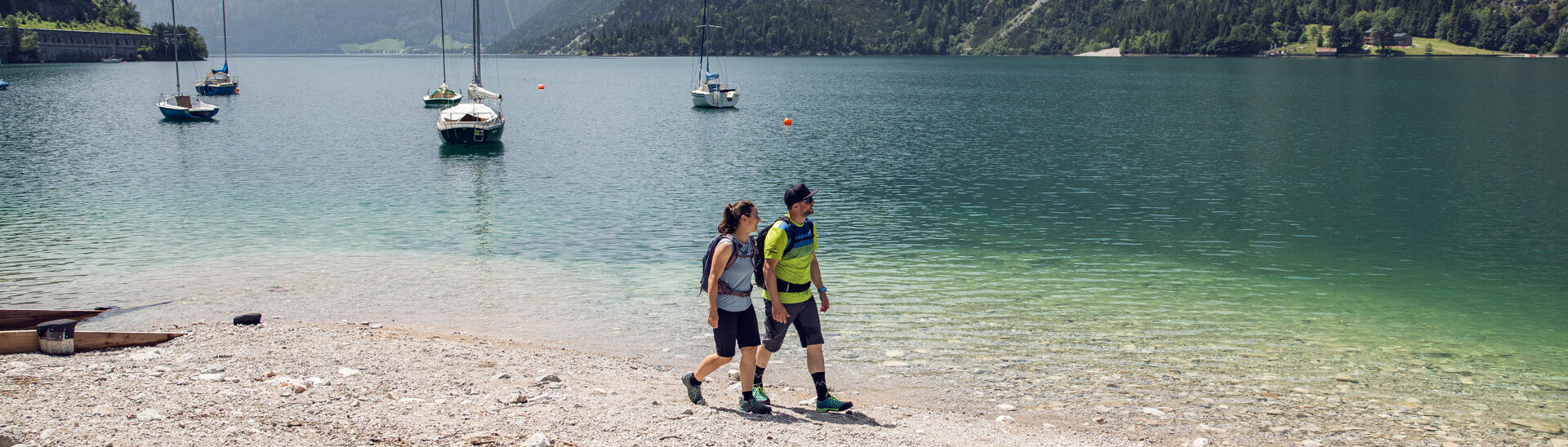 Ein Paar genießt einen kleinen Spaziergang am Ufer des Achensees in Achenkirch. Im Hintergrund sieht man Segelboote.}