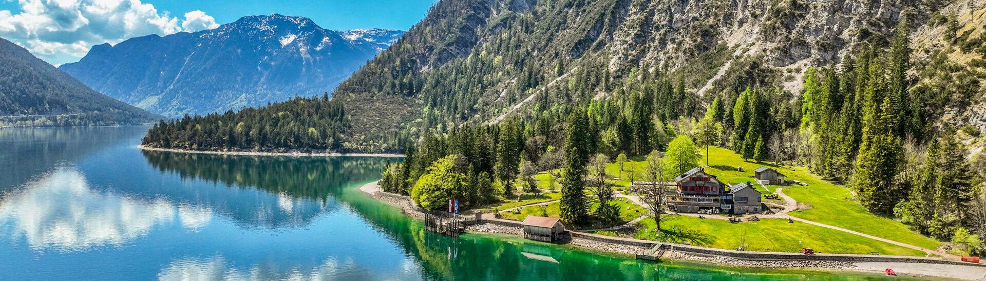 Gaisalm in der Region Achensee Der Achensee umgeben von hohen Bergen und dichtem Wald. Am Ufer steht die Gaisalm, umgeben von Wiesen. Die blau-weißen Wolken spiegeln sich im Wasser, während die Natur in frischem Grün erblüht.}