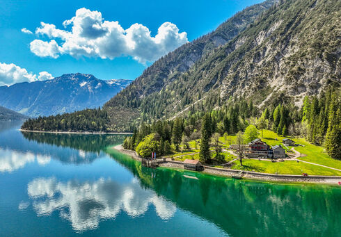Der Achensee umgeben von hohen Bergen und dichtem Wald. Am Ufer steht die Gaisalm, umgeben von Wiesen. Die blau-weißen Wolken spiegeln sich im Wasser, während die Natur in frischem Grün erblüht.