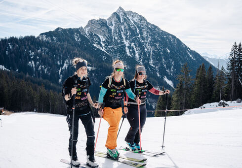 Drei Frauen in Skibekleidung stehen auf einer präparierten Piste im Rofangebirge. Sie lächeln und halten Skistöcke. Im Hintergrund sind majestätische Berge und Bäume sichtbar. Die Stimmung ist fröhlich und sportlich, ideal für einen Skiausflug.