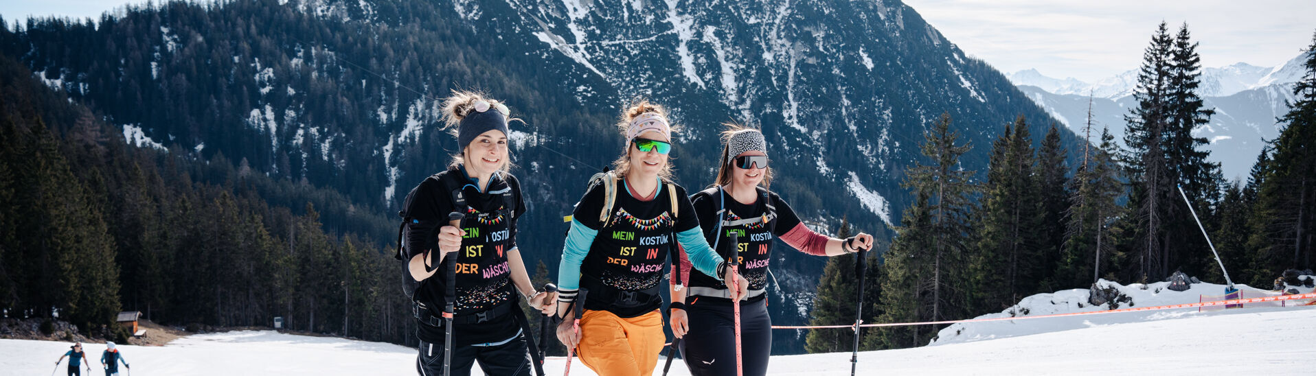 Achensee Xtreme Skitourenspaß Drei Frauen in Skibekleidung stehen auf einer präparierten Piste im Rofangebirge. Sie lächeln und halten Skistöcke. Im Hintergrund sind majestätische Berge und Bäume sichtbar. Die Stimmung ist fröhlich und sportlich, ideal für einen Skiausflug.}