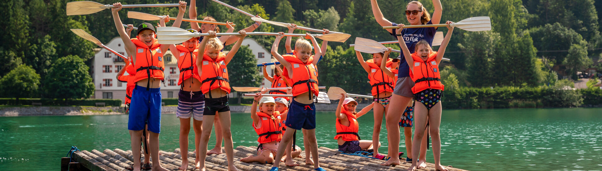 Achensee Kinderprogramm Eine Gruppe von Kindern und einem Erwachsenen steht auf einem Floß im Wasser des Achensees. Sie tragen leuchtend orangefarbene Schwimmwesten und halten Paddel in die Luft. Alle haben fröhliche Gesichtsausdrücke und scheinen Spaß zu haben, während sie gemeinsam posieren.}
