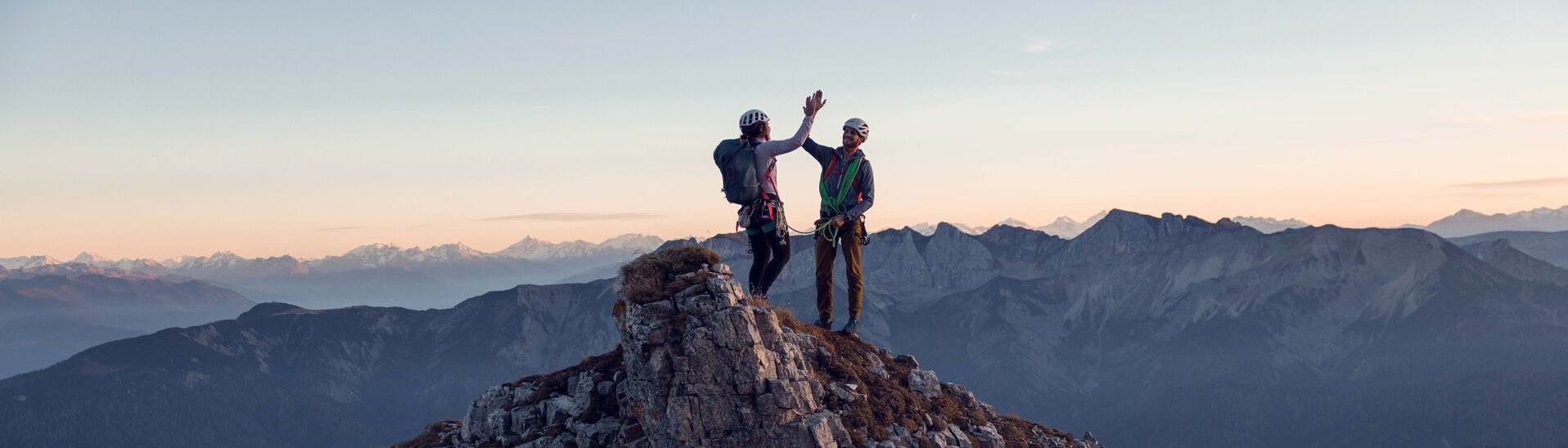 Zwei Kletterer klatschen miteinander am Gipfel des Gufferts bei leicht gerötetem Himmel ein.}