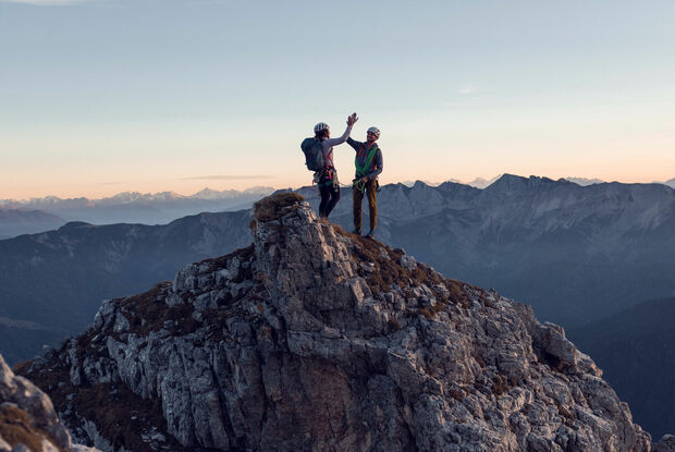 Zwei Kletterer klatschen miteinander am Gipfel des Gufferts bei leicht gerötetem Himmel ein.
