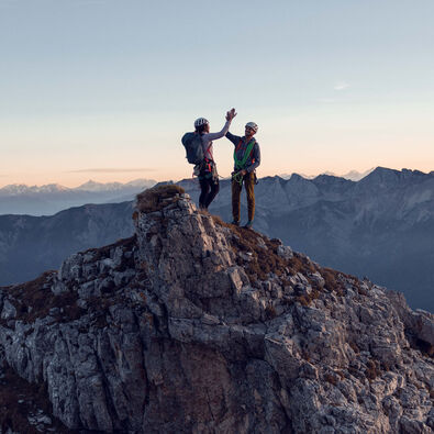 Zwei Kletterer klatschen miteinander am Gipfel des Gufferts bei leicht gerötetem Himmel ein.