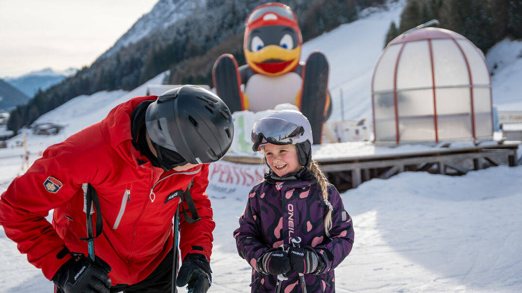 Skifahren bei den Planberg- & Wiesenliften Ein Skilehrer erkundigt sich nach dem Wohlbefinden der kleinen Kursteilnehmerin bei den Planberg- & Wiesenliften in Pertisau. Im Hintergrund Pinguin BOBO.